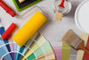 Paint color chips fanned out on a table with a yellow foam paint roller, two paint brushes and paint cans.