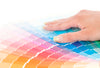 Paint color chips spread out showing shades of oranges, reds, purples, blues and greens, with a person's hand on top of it.