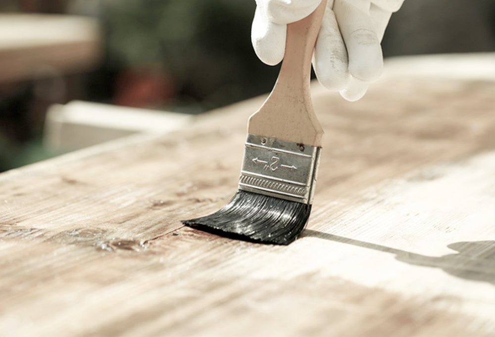 A person's hand wearing a white glove and using a paint brush to put stain on a piece of wood.