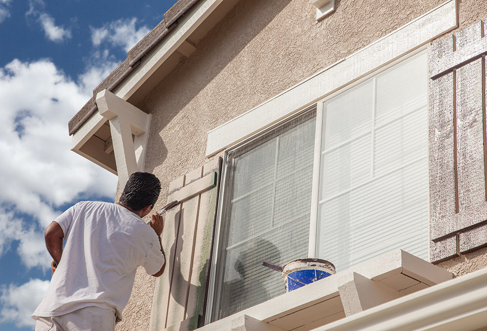 A man wearing all white, painting exterior window shutters on a home.