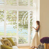 A woman standing by large windows with white Hunter Douglas Palm Beach window shutters, speaking to her phone to use the PowerView motorization app.