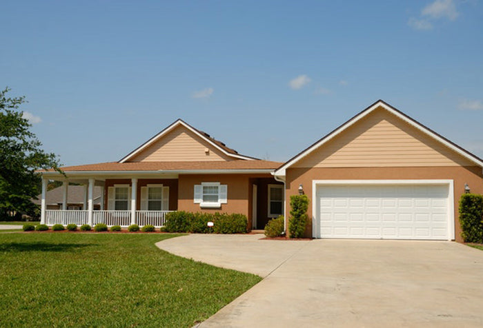 Front view of a light brown house with a white garage and a white porch, with a long front lawn and driveway.