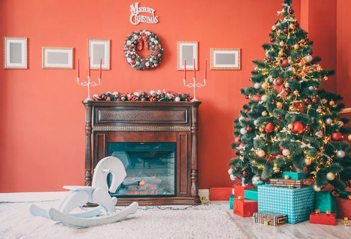 A decorated christmas tree with presents underneath, beside a wooden fireplace with a wreath on top, in front of a red painted wall.