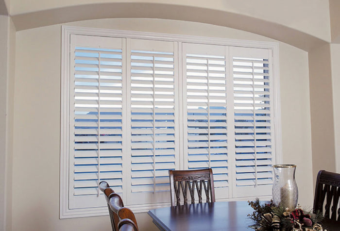 A dining room window with white shutter blinds, with a dark wooden dining room table and chairs.
