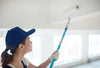 A woman wearing a dark blue ballcap, using a long paint roller to paint a ceiling white.