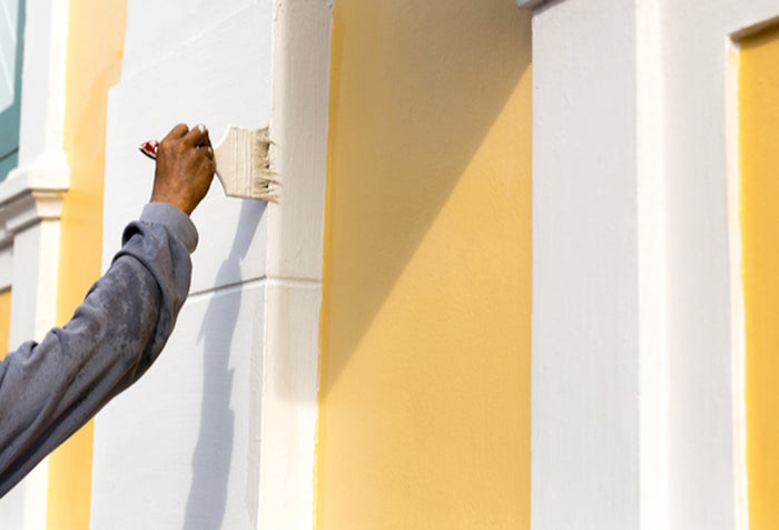 A person's hand panting the exterior of a white and yellow house.