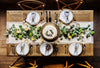 Aerial view of a wooden dining room table with green leaves and white and gold plates and a frosted cake. 
