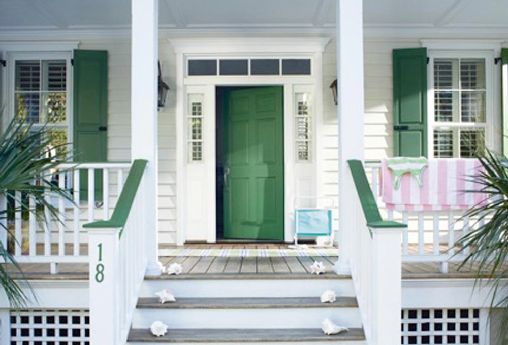 View of the front of a house with a white porch, a green door and green window shutters.
