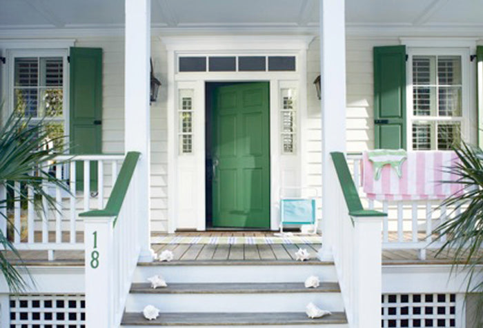 View of the front of a house with a white porch, a green door and green window shutters.