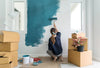 A woman painting a white wall a deep blue teal color, with carboard boxes around her.