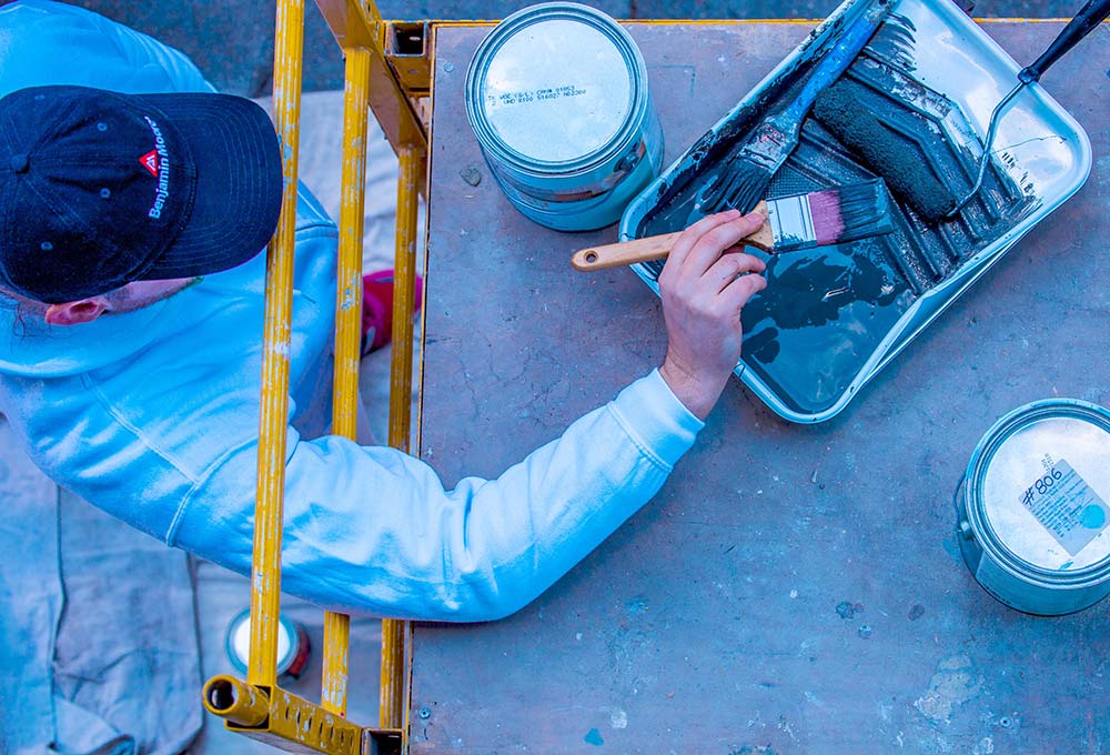 A Painter wearing a Benjamin Moore paints hat, holding a paint brush that he dipped into a paint tray with dark Benjamin Moore paint.