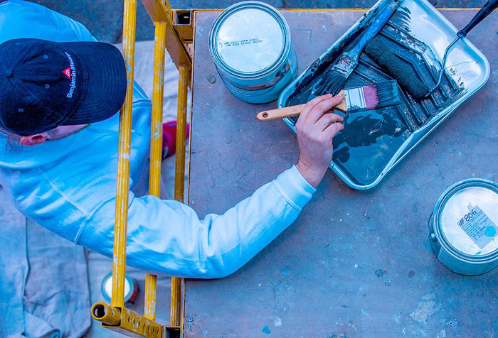 A Painter wearing a Benjamin Moore paints hat, holding a paint brush that he dipped into a paint tray with dark Benjamin Moore paint.