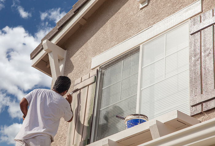 A man wearing all white, painting exterior window shutters on a home.
