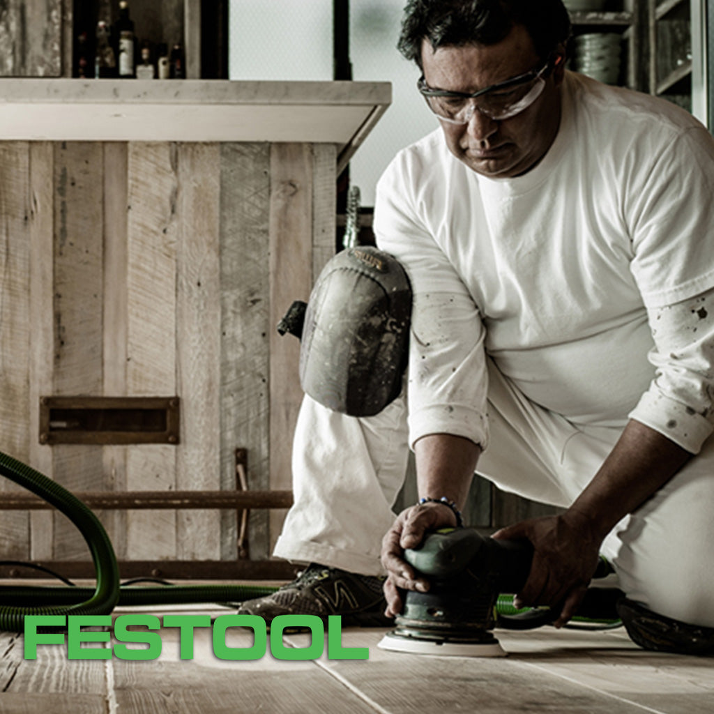 A painter wearing a white painter's outfit and safety glasses, using a Festool abrasive tool on a hardwood floor.