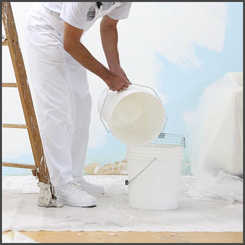 A man wearing white painter's overalls, pouring primer from one white pail into another pail, standing in front of a half-primed wall.